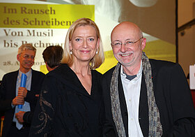 Zwei Personen in eleganter Kleidung lächelnd vor einer Leinwand. 