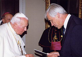 Weißhaarige Person im schwarzen Anzug überreicht Papst Johannes Paul II eine Urkunde.