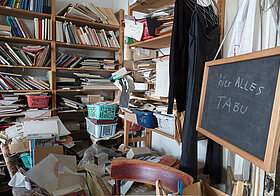 Foto von chaotischem Raum mit vollen Bücherregalen, rechts eine Kreidetafel mit Aufschrift "Hier alles tabu"