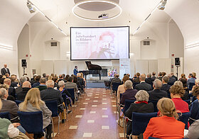 Saal mit Sesselreisen und Publikum, eine Leinwand hängt über der Bühne.