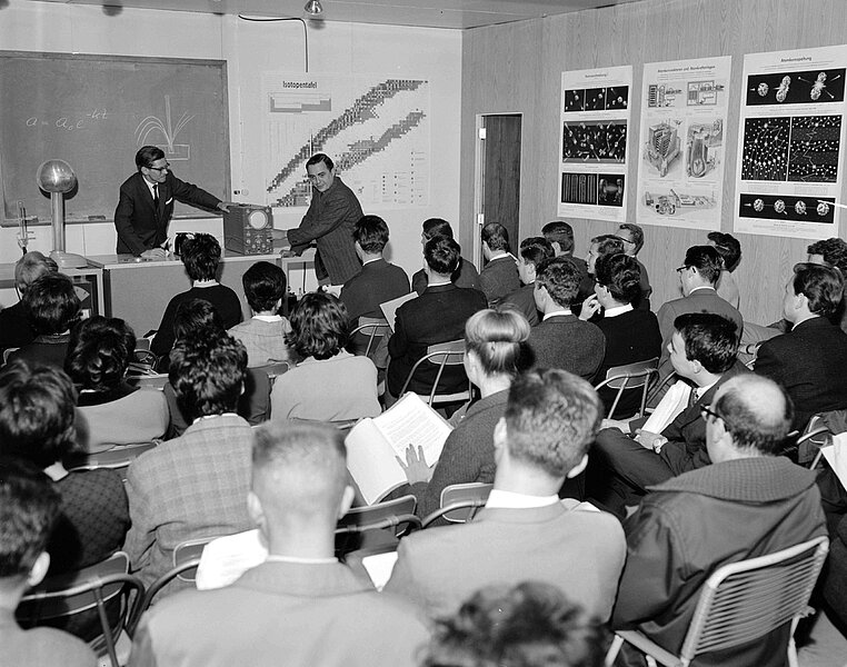 Ein Klassenraum mit Tafel voller Menschen, physikalische Poster an den Wänden, schwarz-weiß