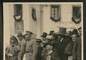 Schwarz-weißes Foto von Person in Tracht mit Ziehharmonika, daneben Personen in Uniform.