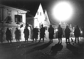 Eine Reihe an Personen mit Helmen geht nebeneinander auf einer Straße, sie sind komplett im Schatten, die Häuser dahinter sind beleuchtet.