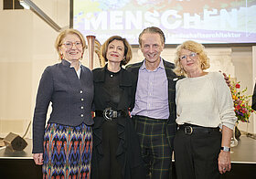 Vier elegant gekleidete Personen posieren lächelnd vor einer Bühne. Im Hintergrund eine Leinwand. 