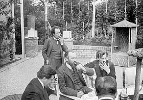 Fünf Personen in Anzügen sitzen auf einer Terrasse und rauchen.