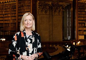 Porträtfoto von einer blonden Frau in dunkler, gemusterter Bluse vor Bücherregalen