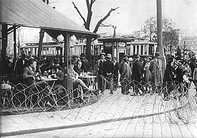 Personen sitzen in Café draußen und stehen bei einem Kiosk, im Vordergrund Stacheldrahtzaun.