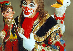 Ein Clown mit einer Kasperlpuppe und einer Stofftiergans.