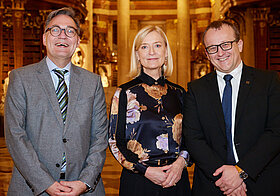 Zwei Personen im Anzug und eine elegant gekleidete Person im Prunksaal.