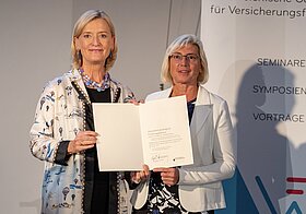 Zwei Personen in eleganter Kleidung halten ein Dokument vor einer Leinwand. 