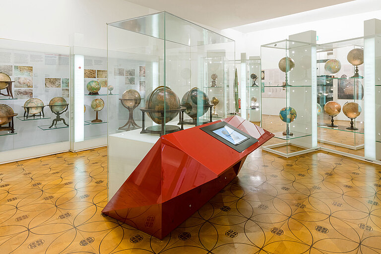 Museumsraum mit Globen in Glasvitrinen und einem großen, roten Tisch mit Bildschirmen.