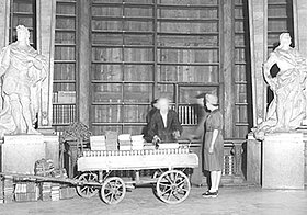 Zwei Personen mit einem Wagen voller Bücher vor leeren Bücherregalen