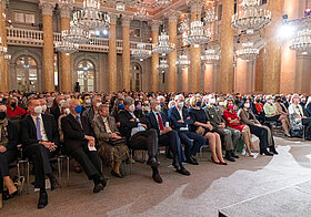 Volles Publikum in einem Marmorsaal.