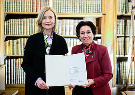 Zwei Personen nebeneinander halten zusammen eine Urkunde, im Hintergrund ein Bücherregal.