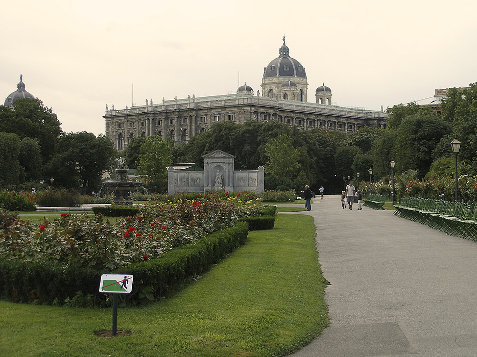 Foto eines großen Stadtparks mit Rosen und Parkbänken und einem großen Museumsgebäude im Hintergrund.