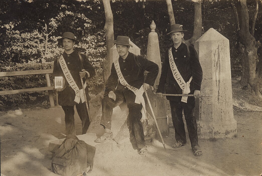 Drei Personen nebeneinander, schwarz-weißes Foto. Alle tragen Anzüge, Hüte, Schärpen und Sandalen. Einer von ihnen hält ein Foto der Gruppe in der Hand. Hinter ihnen Wald, ein Zaun und Stein-Skulpturen.