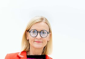 Porträtfoto von einer blonden Frau in orangeroter Jacke und schwarz umrandeter Brille.