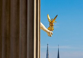Blick durch Fenster auf alte Gebäude mit Kirchtürmen und rot-weiß-roter Flagge, seitlich eine Statuen-Hand, die eine kleinere, goldene Hand hält.