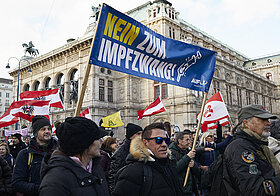 Personen auf Demozug mit rot-weiß-roten Flaggen und einem Banner, Text: "Nein zur Impfpflicht".