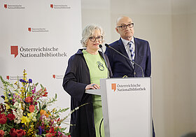 Person mit grünem Oberteil am Rednerpult, daneben Person in blauem Anzug.