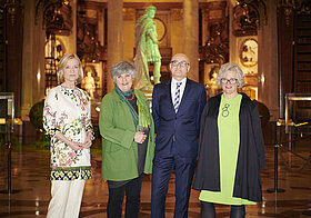 Vier elegant gekleidete Personen in einem alten Bibliothekssaal mit grün beleuchteter Statue im Hintergrund.