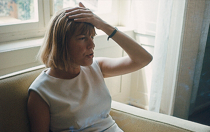 Foto von Ingeborg Bachmann, die auf einem Sofa sitzt und sich an den Kopf greift. 