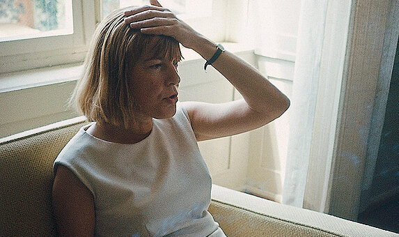 Foto von Ingeborg Bachmann, die auf einem Sofa sitzt und sich an den Kopf greift. 