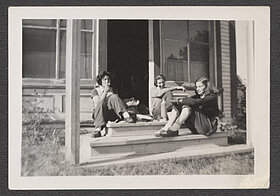 Schwarz-weißes Foto von drei jungen Menschen, die auf einer Veranda sitzen. Eine von ihnen raucht eine Zigarette.