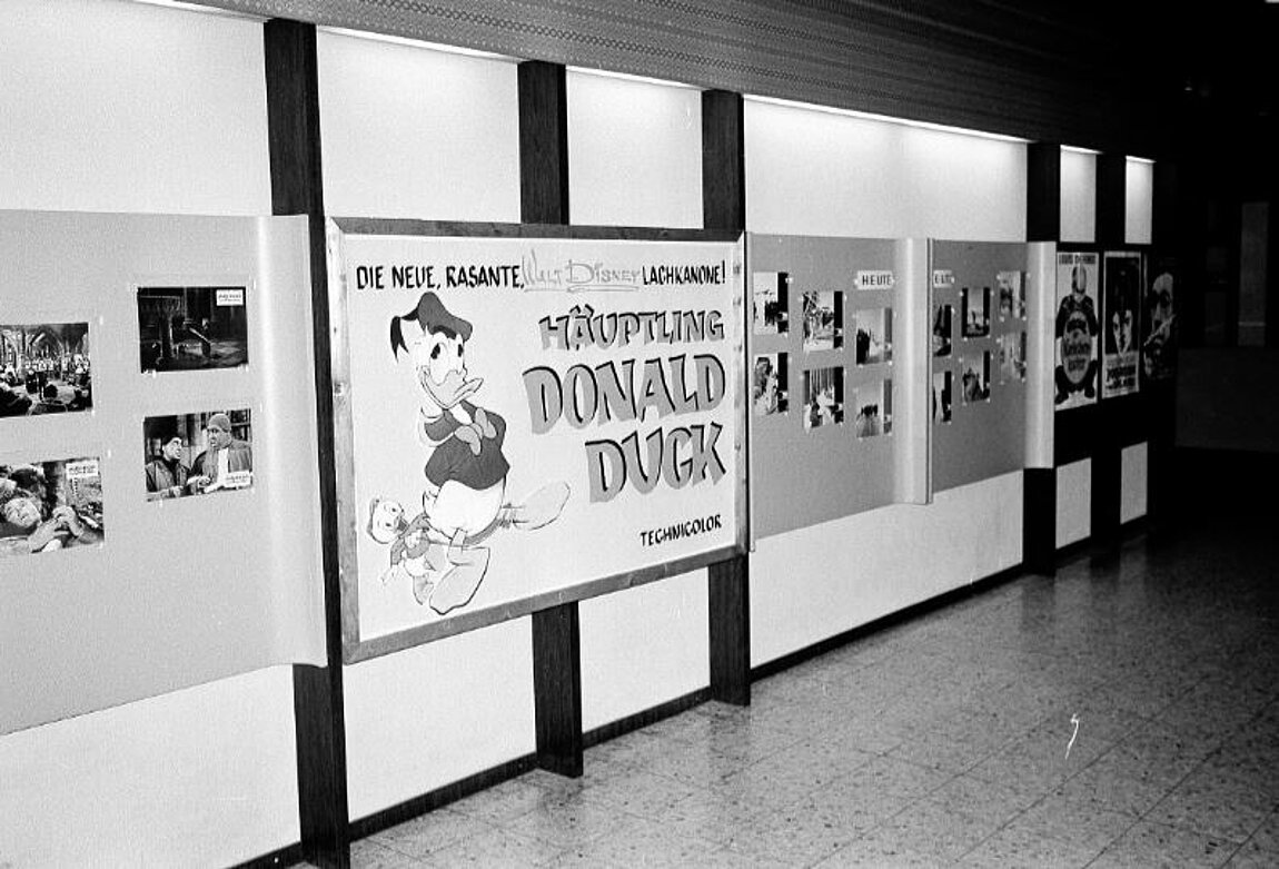 Schwarz-weißes Foto von Wand, auf der viele Fotos und ein Donald Duck Plakat angebracht sind