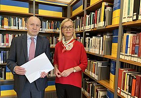 Zwei elegant gekleidete Personen posieren vor Bücherregal mit ukrainischer Flagge. 
