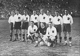 Fußballmannschaft auf Rasen in Stadion in weißen, langärmligen Oberteilen mit Wappen.