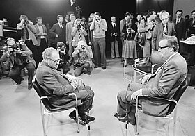 Schwarz-Weiß-Foto von einem Raum mit mehreren Menschen, zwei Menschen zuhören, die auf einem Stuhl sitzen und reden.