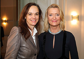 Eine dunkelhaarige und eine blonde Person lächeln in die Kamera.