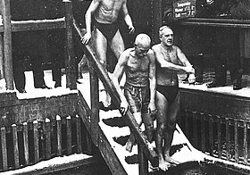 Männer in Badehosen auf einer Stiege, dahinter Männer in Wintermänteln