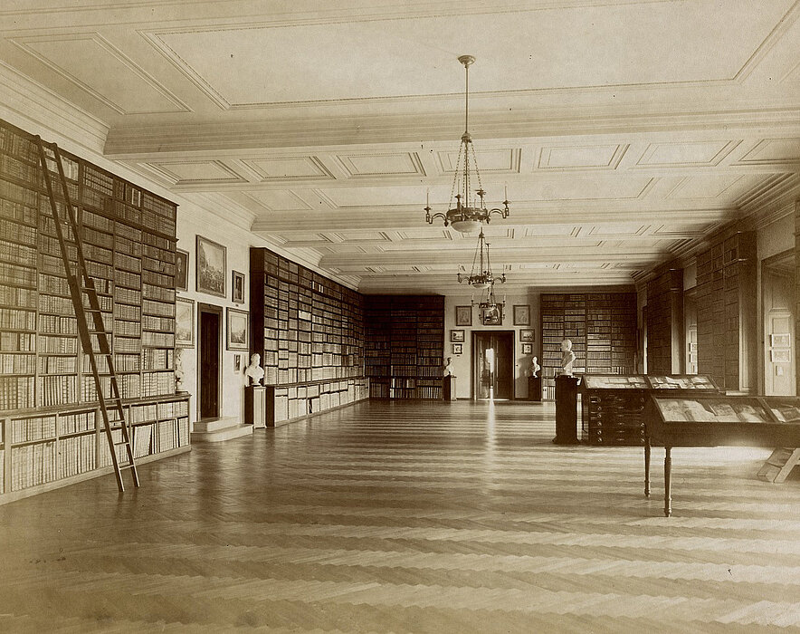 Saal mit großen Bücherregalen, Büsten und Kronleuchtern, Schwarz-Weiß-Fotografie.