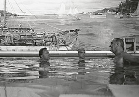Drei Personen schwimmen in einem Indoor-Pool, im Hintergrund ein Gemälde mit Schiffen.