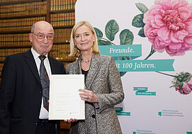 Zwei Personen halten eine Urkunde vor einer Logowand und einem Bücherregal.