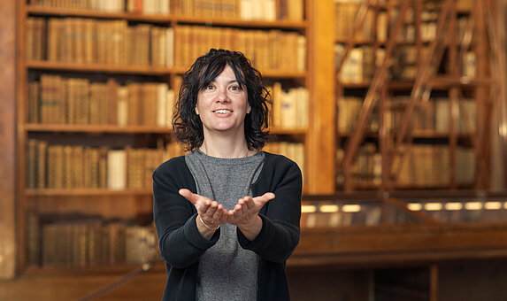 Foto von Person vor einer Vitrine und Bücherregalen, die "Buch" in Gebärdensprache zeigt.