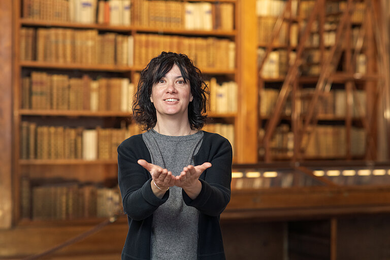 Foto von Person vor einer Vitrine und Bücherregalen, die "Buch" in Gebärdensprache zeigt.
