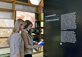 Dauerausstellung des Literaturmuseums. Zwei Frauen verwenden das Medientablet, um zusätzliche Informationen über die Objekte abrufen zu können.