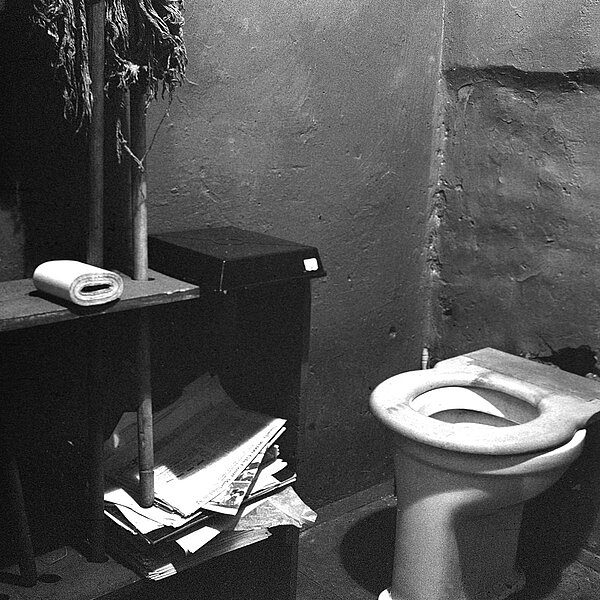 Karge Toilette, daneben Zeitungen und Klopapier, schwarz-weiß