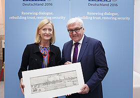 Zwei Personen posieren mit länglicher Zeichnung in weißem Rahmen vor blauen Aufstellern.
