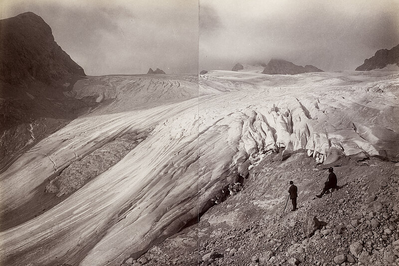 Schwarz-weißes Foto von Eisfläche bei Bergen, rechts zwei Wanderer.