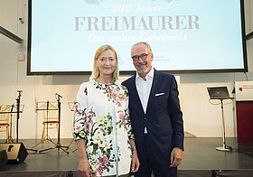 Zwei Personen in eleganter Kleidung posieren vor Leinwand und Bühne.