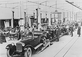 Altmodische Autofabrik, in der Mitte eine Reihe an fertigen Autos, die Arbeiter schauen zur Kamera.