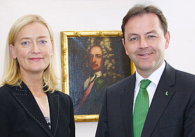 Zwei Personen posieren mit aufgeschlagenem Buch vor einem Portrait. 