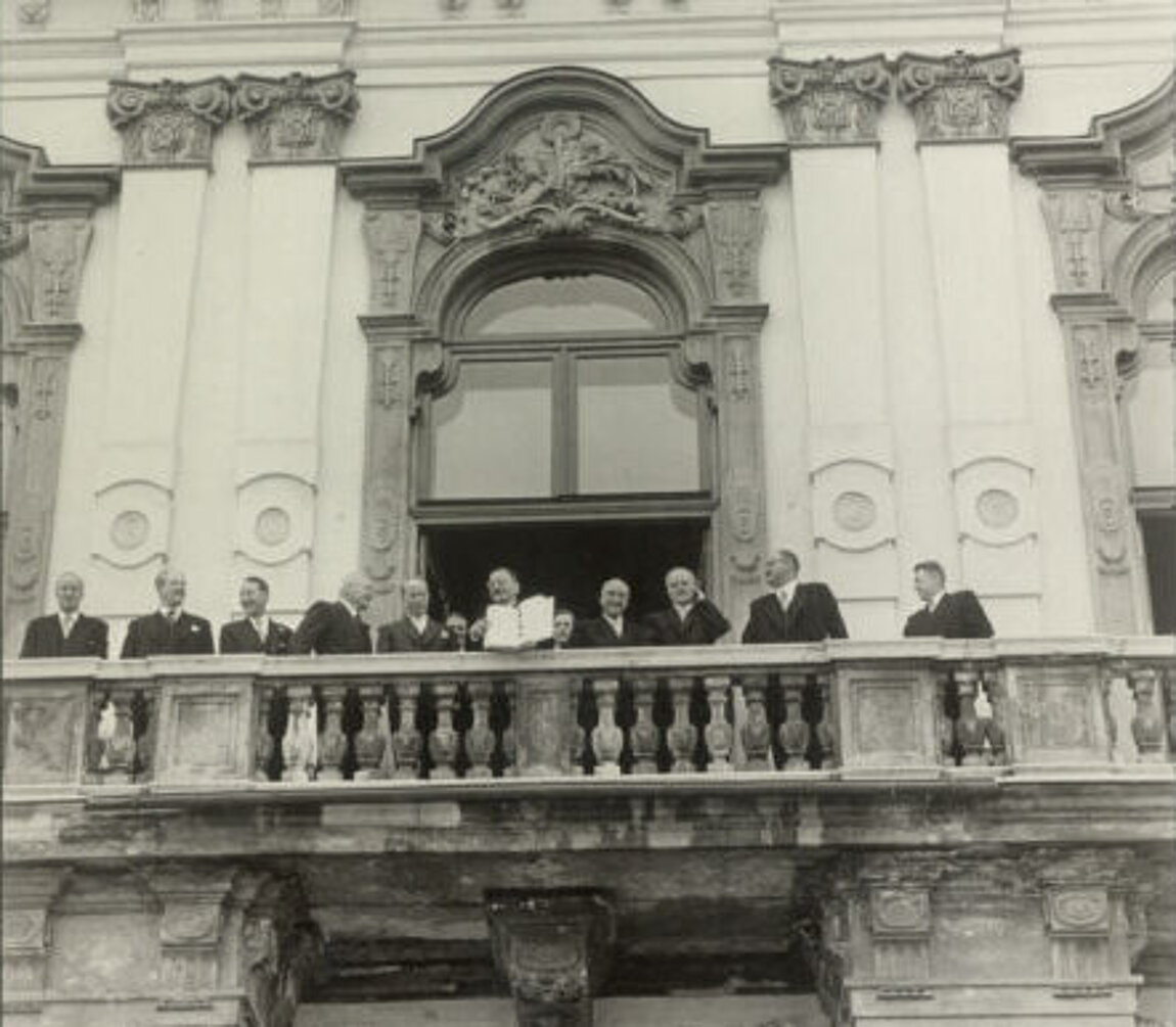 Schwarz-Weiß-Foto eines historischen Gebäudes mit mehreren Menschen, die auf einem Balkon stehen.