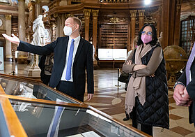 Einige Personen in eleganter Kleidung stehen in einem Marmorsaal vor Vitrinen.