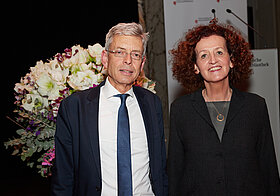 Foto von zwei Personen im Anzug. Im Hintergrund ein Blumenstrauß.