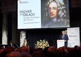Bebrillte Person am Rednerpult vor einer Leinwand. 